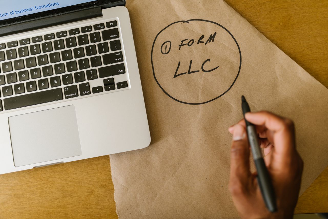 services-04 Close-up of hand writing 'Form LLC' on craft paper beside a laptop, indicating business planning.